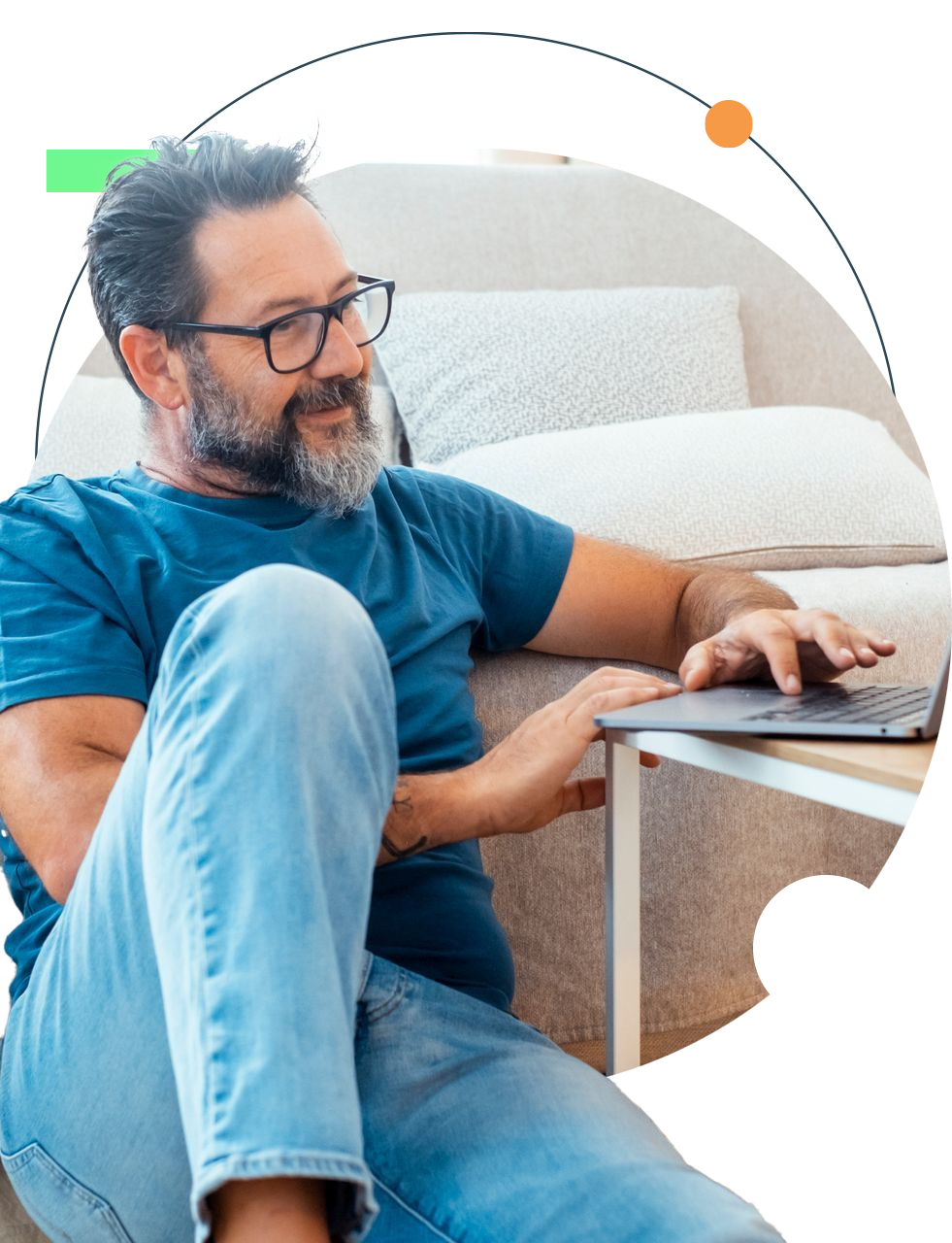 Man in glasses, smiling, using a laptop while seated on the floor near a couch.