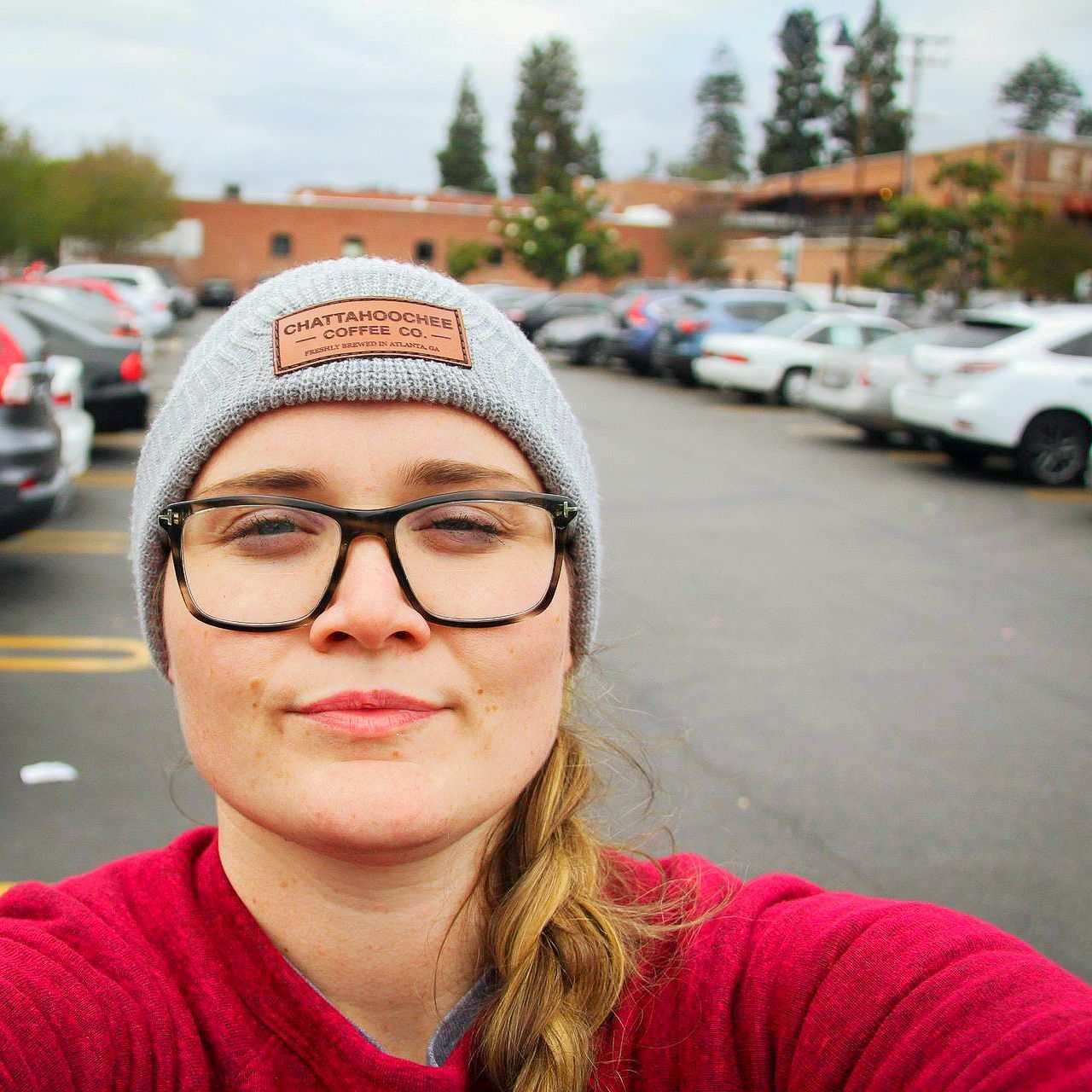 Woman in glasses and beanie smiles in a parking lot, with cars and buildings in the background.