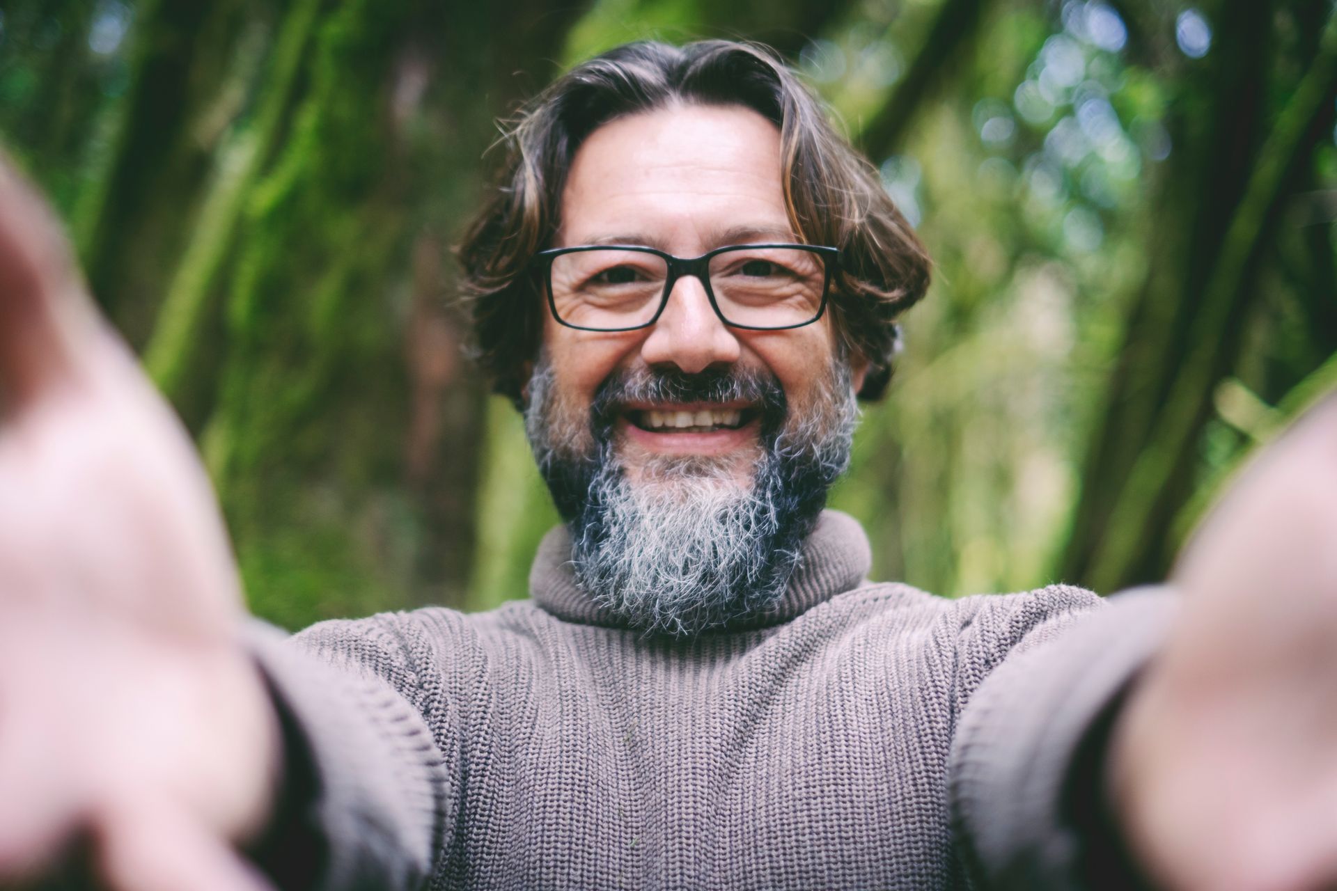 Man with glasses and beard smiling, arms outstretched in forest setting.