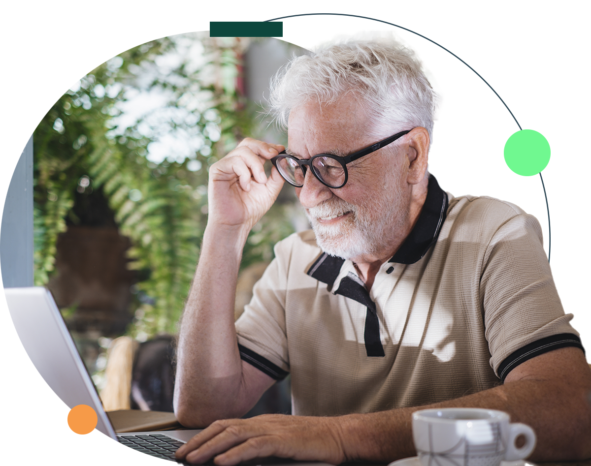 Senior man with glasses using a laptop, smiling. Sitting at a table, indoors, with coffee.