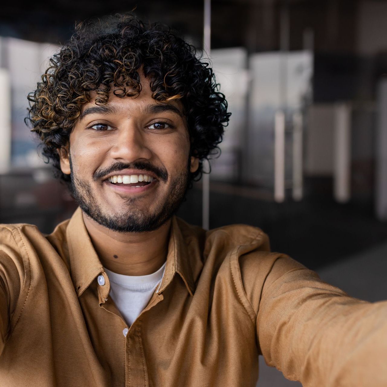 Man with curly hair smiling, taking a selfie in an office setting. He wears a brown shirt.