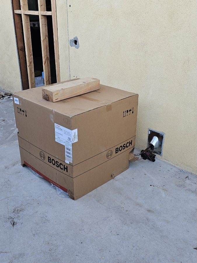 Stacked cardboard boxes with a brick on top, sitting on a concrete floor beside a wall outlet.