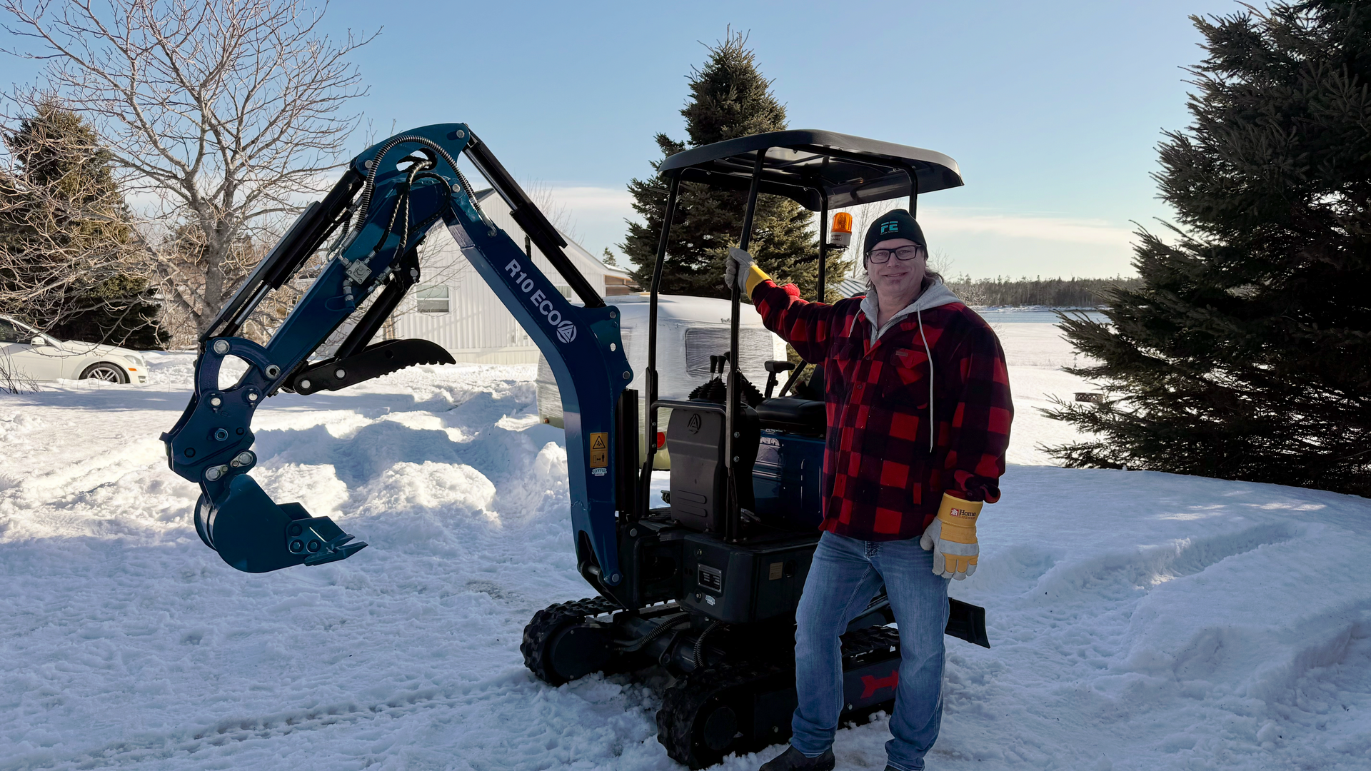 A person in a plaid jacket stands next to a blue Rippa mini-excavator in a snowy, Nova Scotai winter after receiving a delivery from Joyful Group.