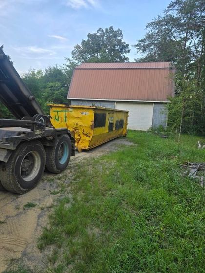 Yellow dumpster being loaded by truck, parked beside a white building with an orange roof, outdoors.