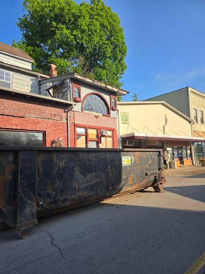 A large dumpster parked on a street in front of a brick building and a row of shops.