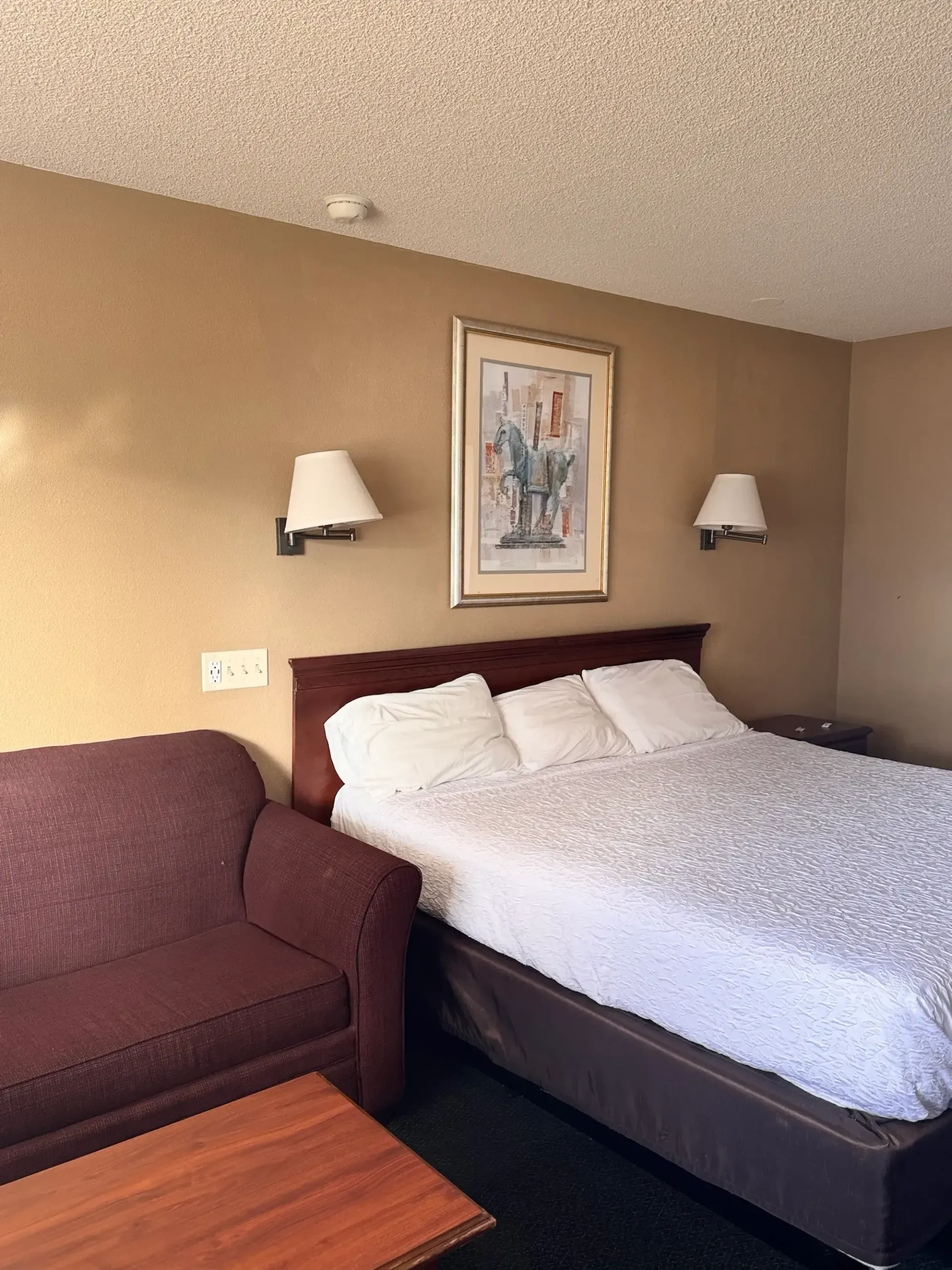 Hotel room with a bed, small sofa, artwork, and lamps on the wall.
