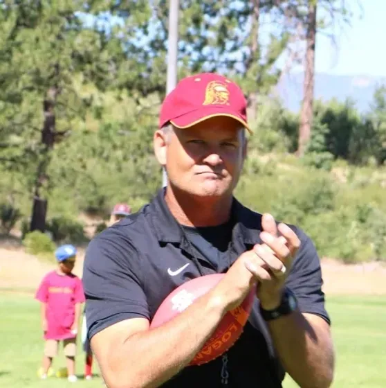 Man with crossed arms on a football field. He is wearing a blue polo shirt, khaki pants, and has a serious expression.