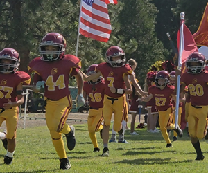 Flag football game at night, a player in red jersey runs with the ball as others pursue on a green field.