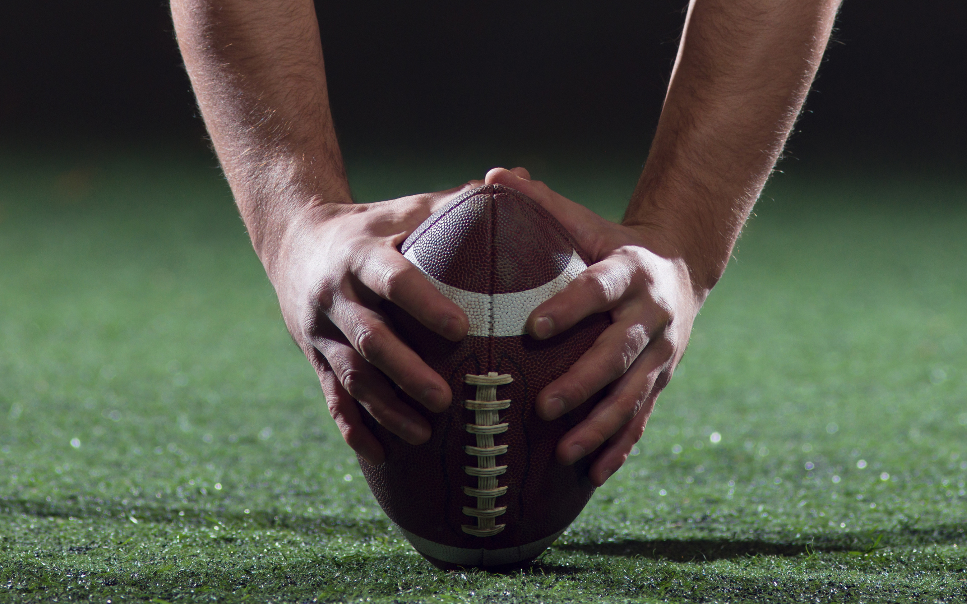 Man in green polo stands on a football field, arms crossed, stadium in background.