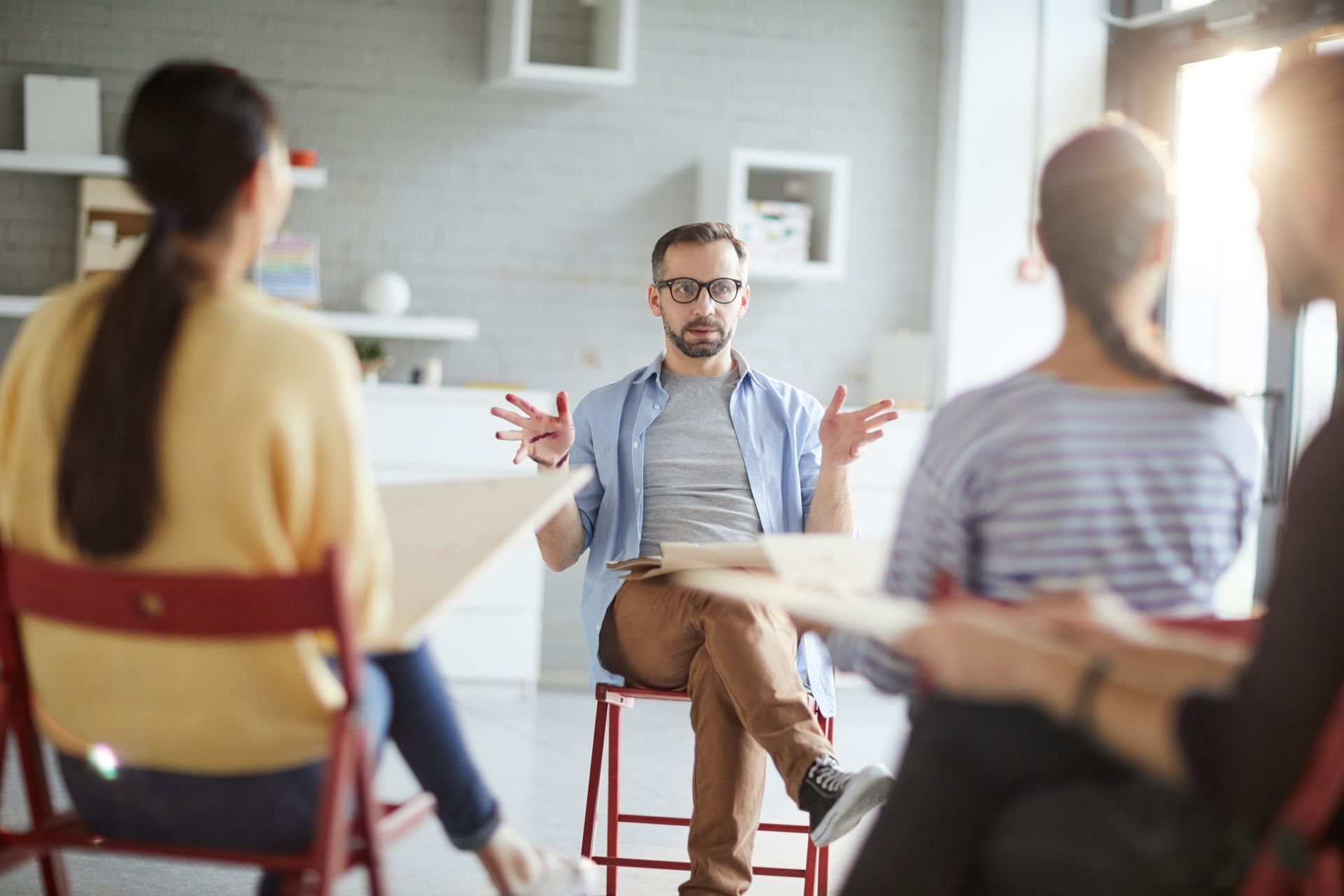 Man in glasses speaking to a small seated group in a bright meeting room with white walls and stools