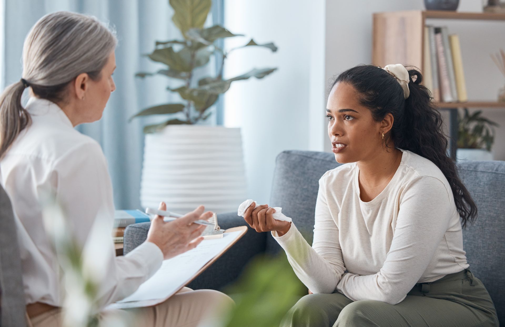 Two women talking in a bright counseling office, one taking notes while the other gestures.