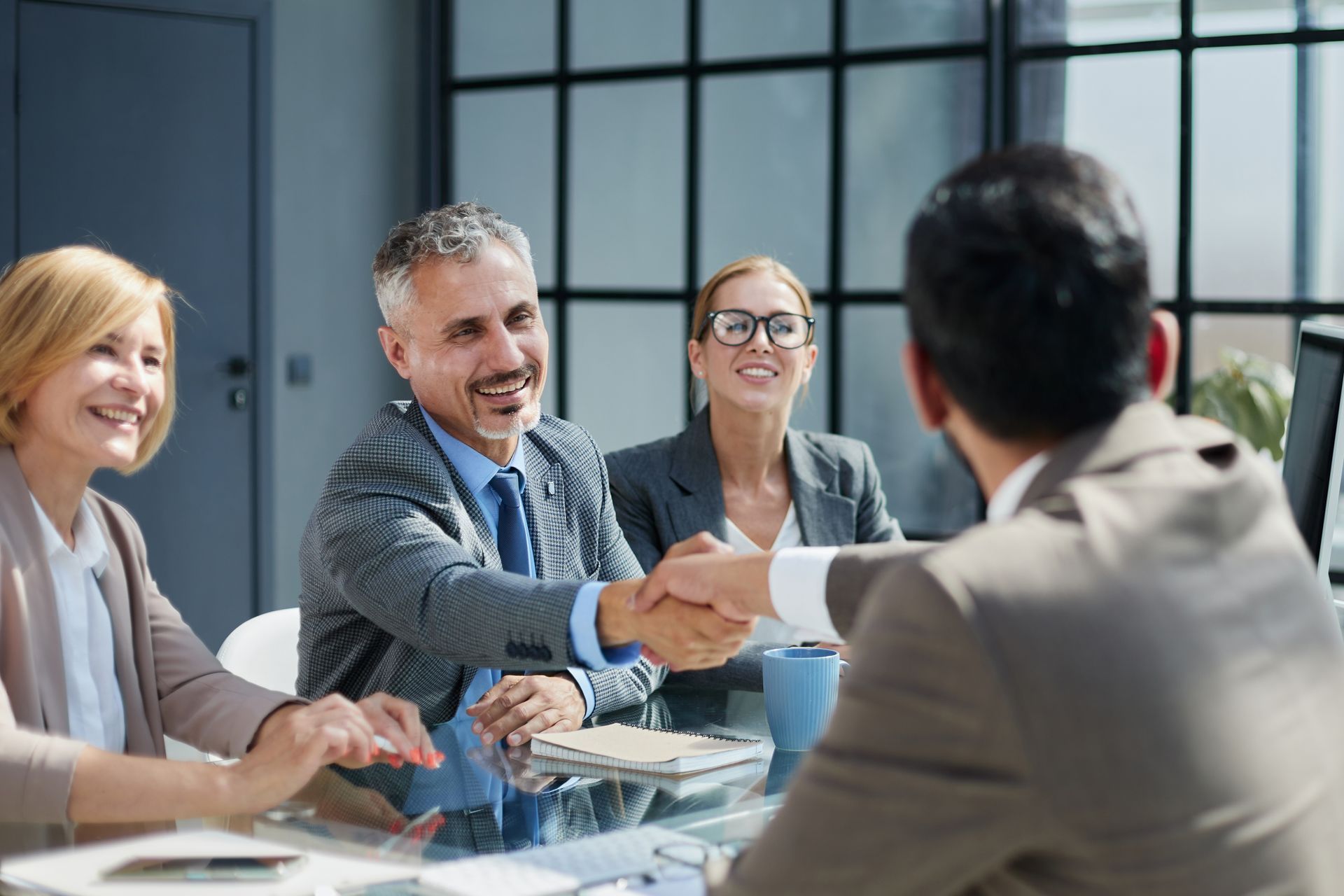 Business meeting with four people around a glass table, smiling and shaking hands in a bright office.