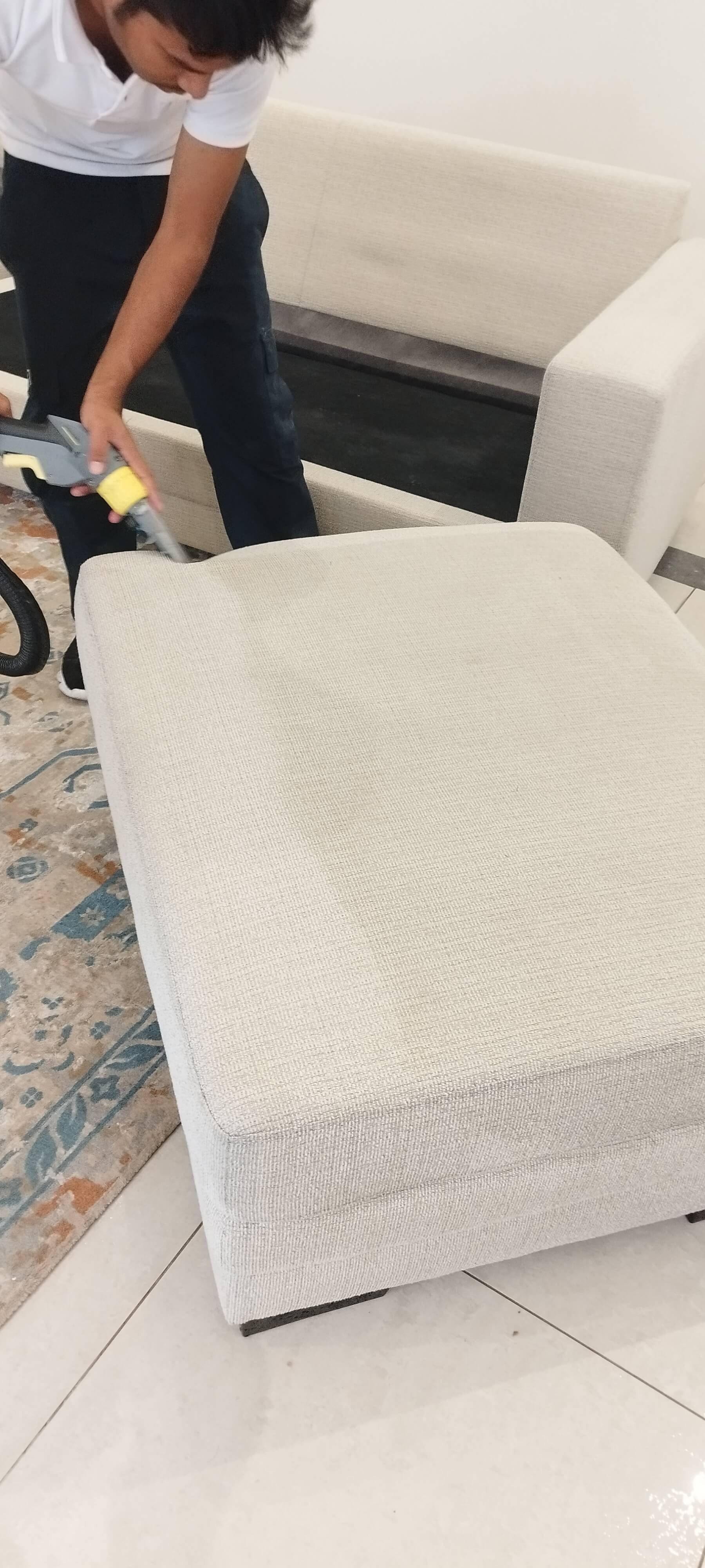 A man is cleaning a white ottoman with a vacuum cleaner.