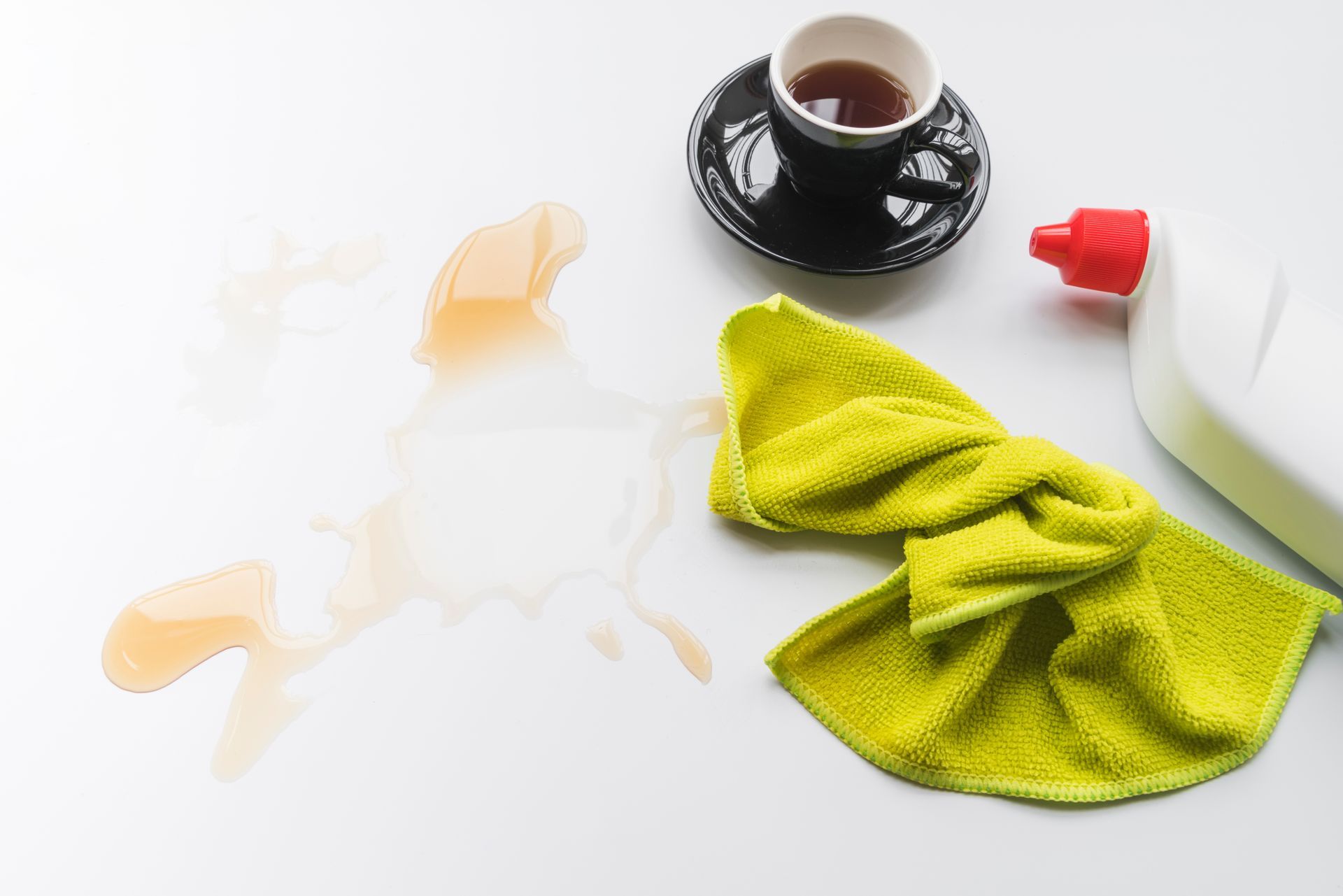 Spilled tea on white surface with a cup, cleaning spray, and yellow cloth.