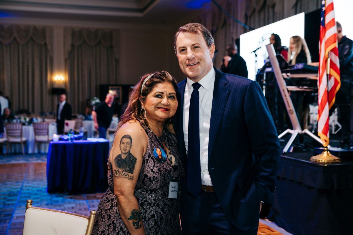 Woman with a tattoo and man smiling at an event, posing for photo near stage with band and US flag.
