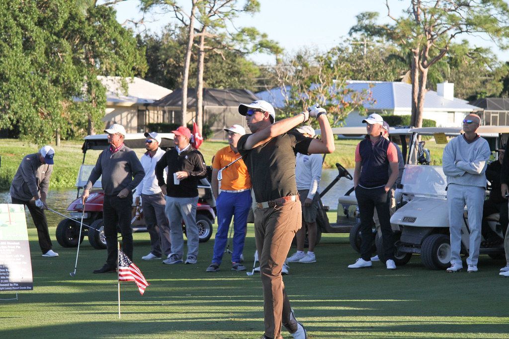 Golfer swings on a course; brown pants, green shirt. People watch, golf carts nearby, sunny day.