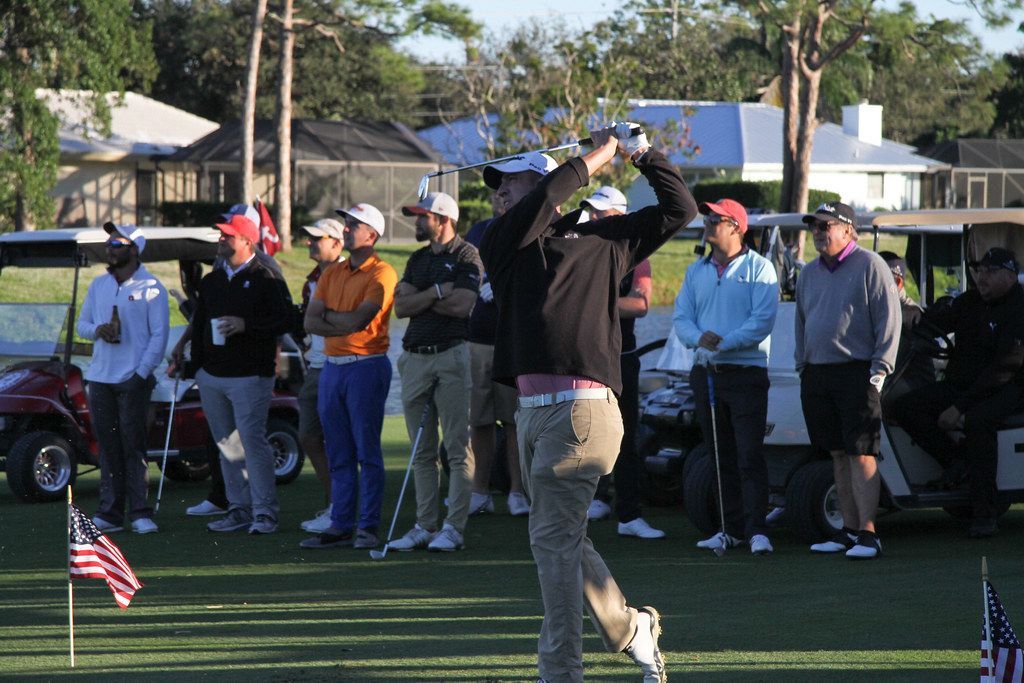 Golfer swinging club on a green with spectators; sunny outdoor setting with golf carts and houses.
