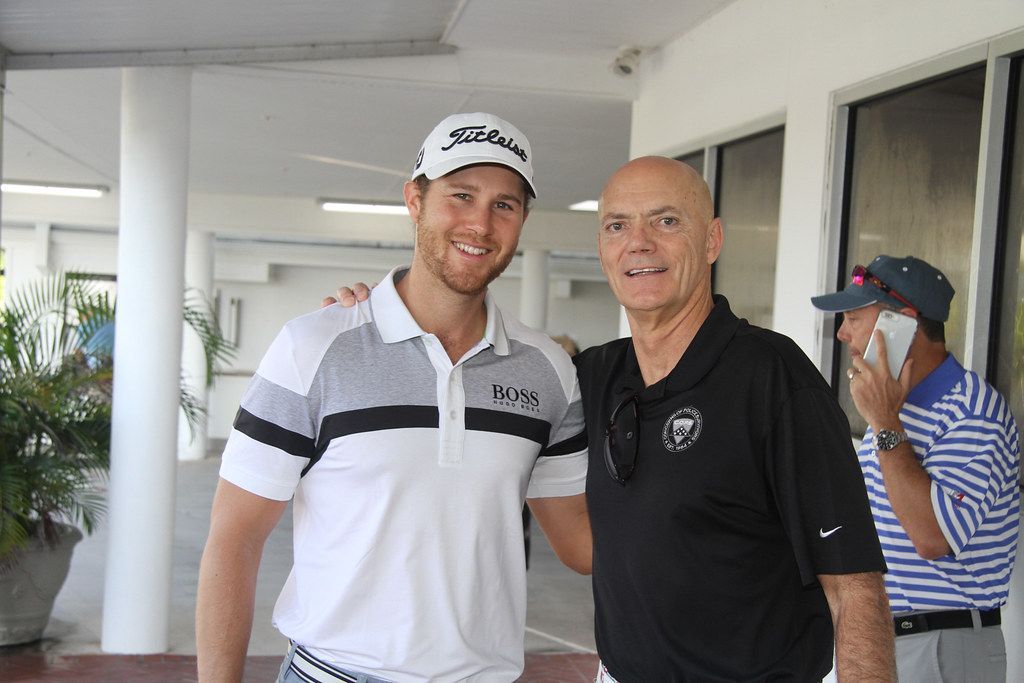 Two men smiling, arm in arm, outdoors; one in golf attire, the other in black polo shirt, another man on phone.