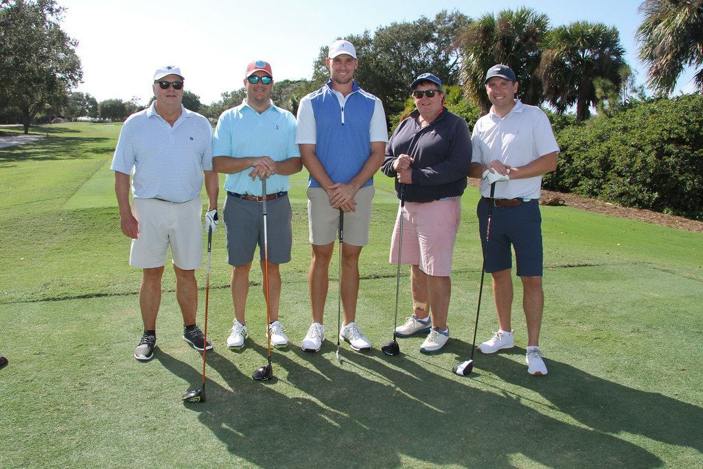 Five men in golf attire pose on a green, sunny day.