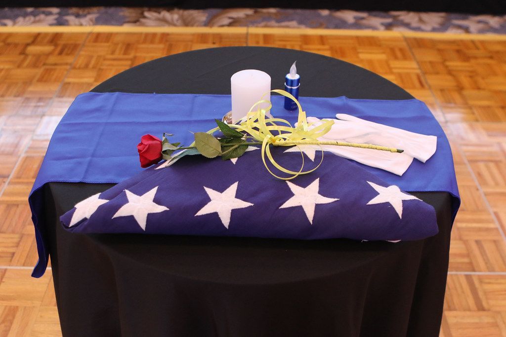 Ceremonial display: folded American flag, red rose, candle, blue runner on a black table in a hall.