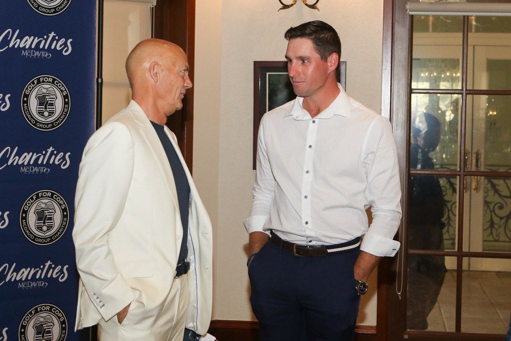 Two men talking indoors: one in a white suit, one in a white shirt and navy pants.