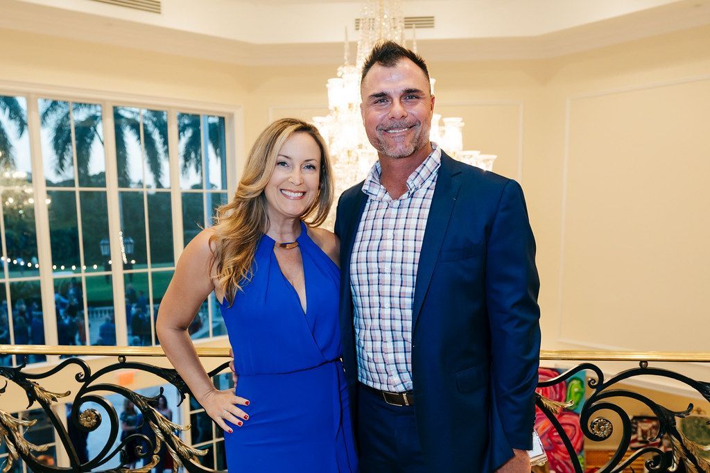 Woman in blue gown and man in blue blazer smiling together, indoors, near a balcony.