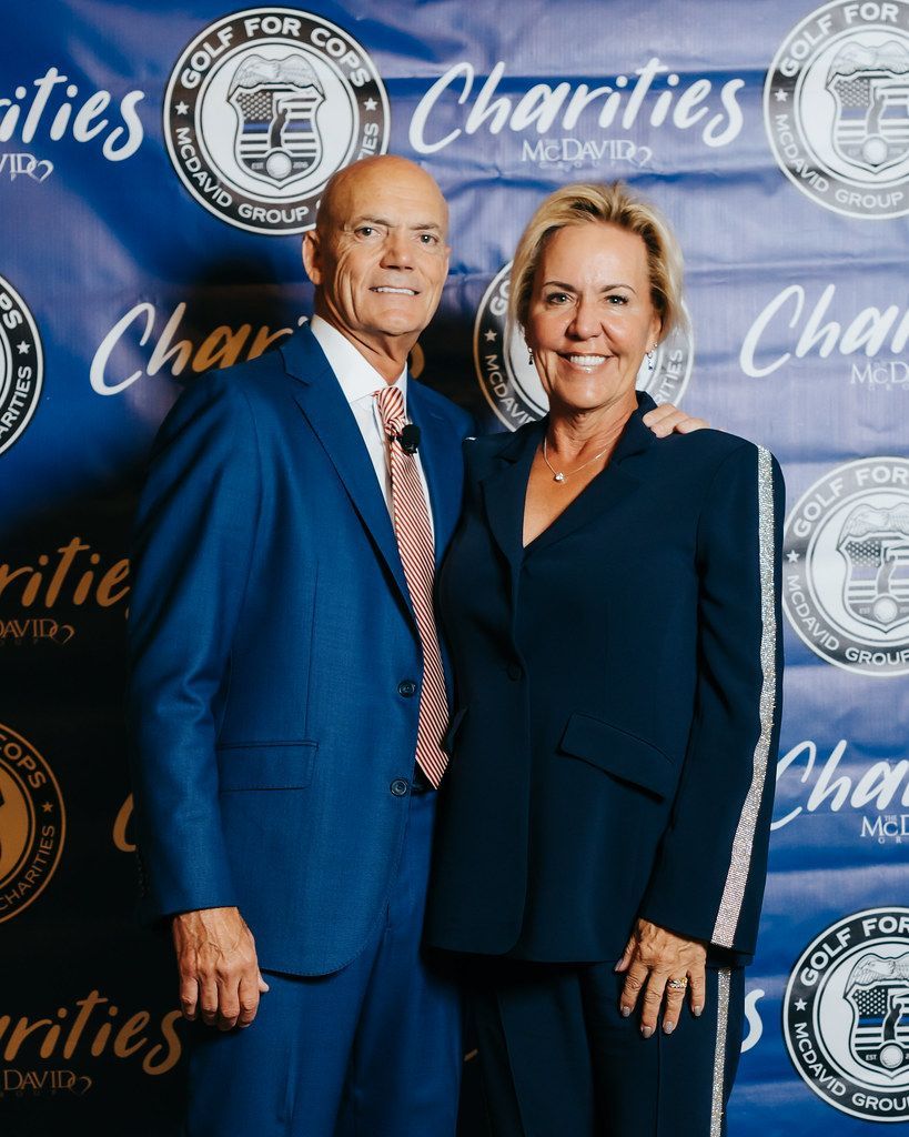 An older couple, smiling, pose in front of a banner. The man wears a blue suit. The woman wears a navy suit.