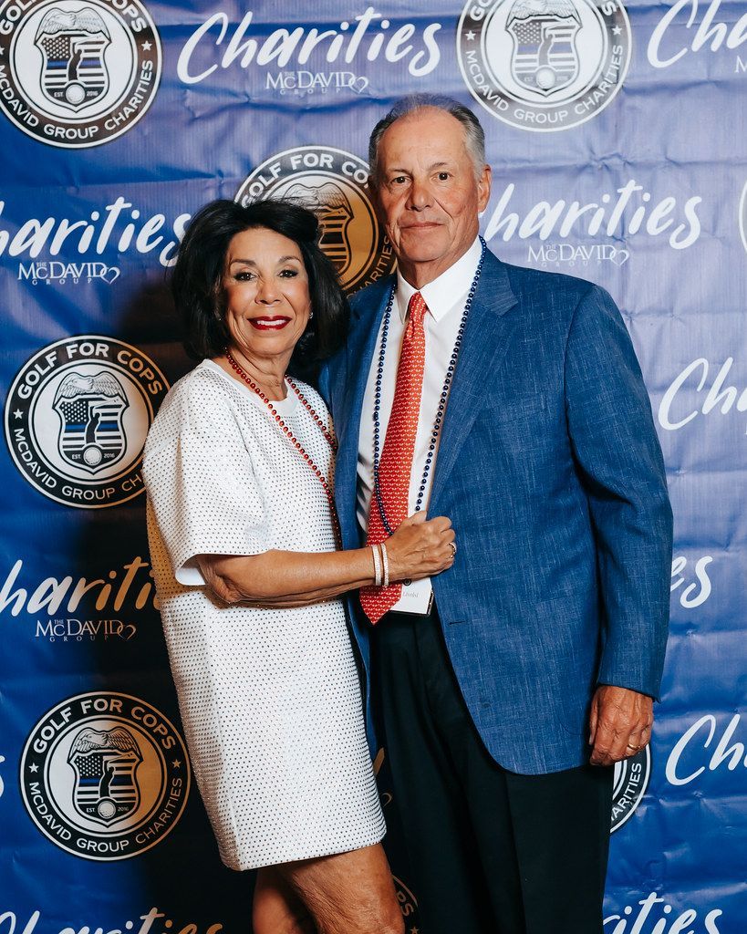 Woman and man pose together; she wears white dress, he wears blazer and red tie, blue backdrop.