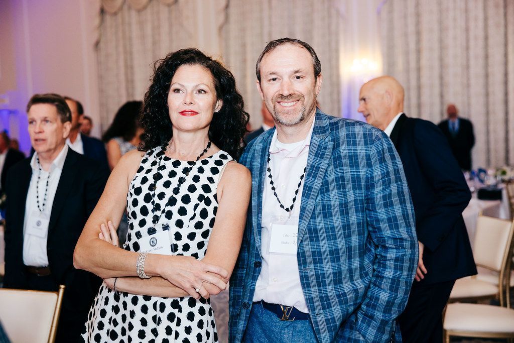 Woman in black and white polka-dot dress and man in plaid jacket pose at an event, indoors.
