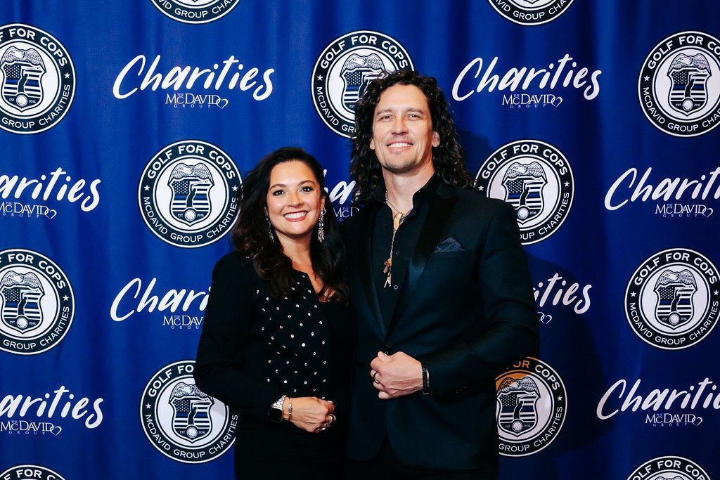 Man and woman pose in front of a blue backdrop with logos. The woman wears black; the man wears a black suit.