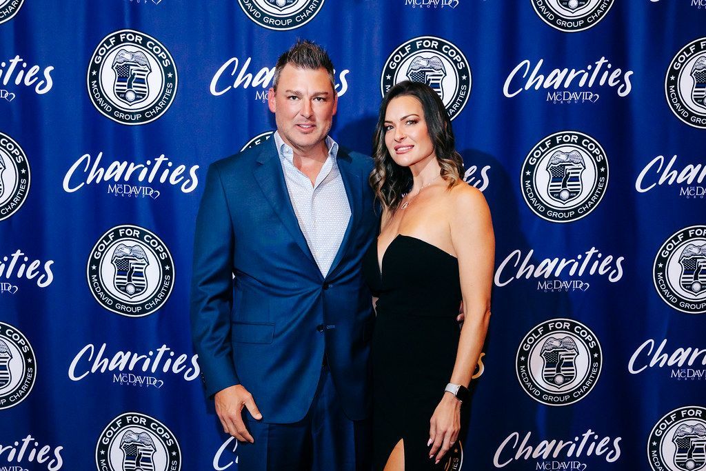 A man and woman pose in front of a blue backdrop with logos. Man wears a blue suit, woman wears a black dress.