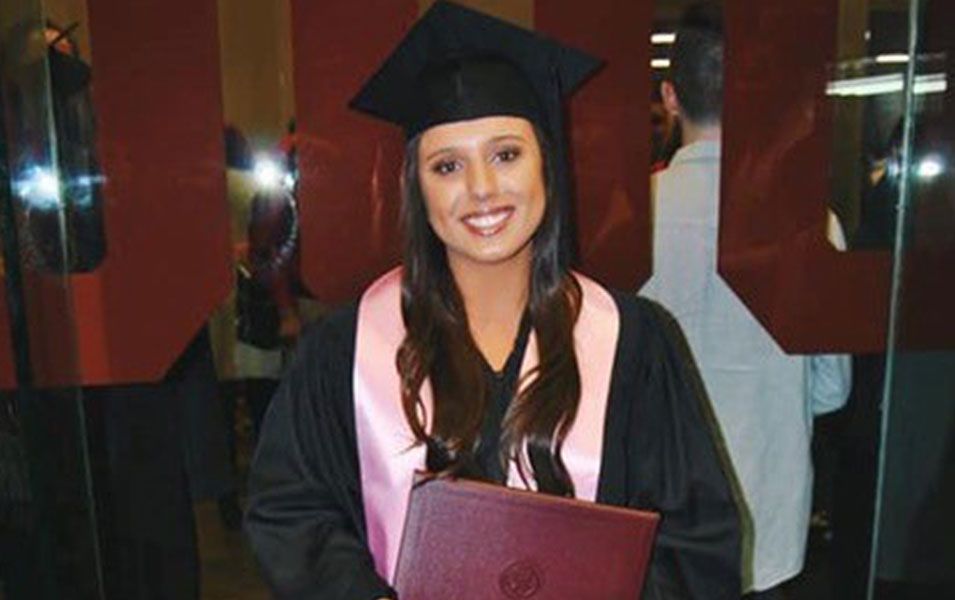 Woman in graduation cap and gown smiles, holding diploma, pink stole, in front of mirrored wall.