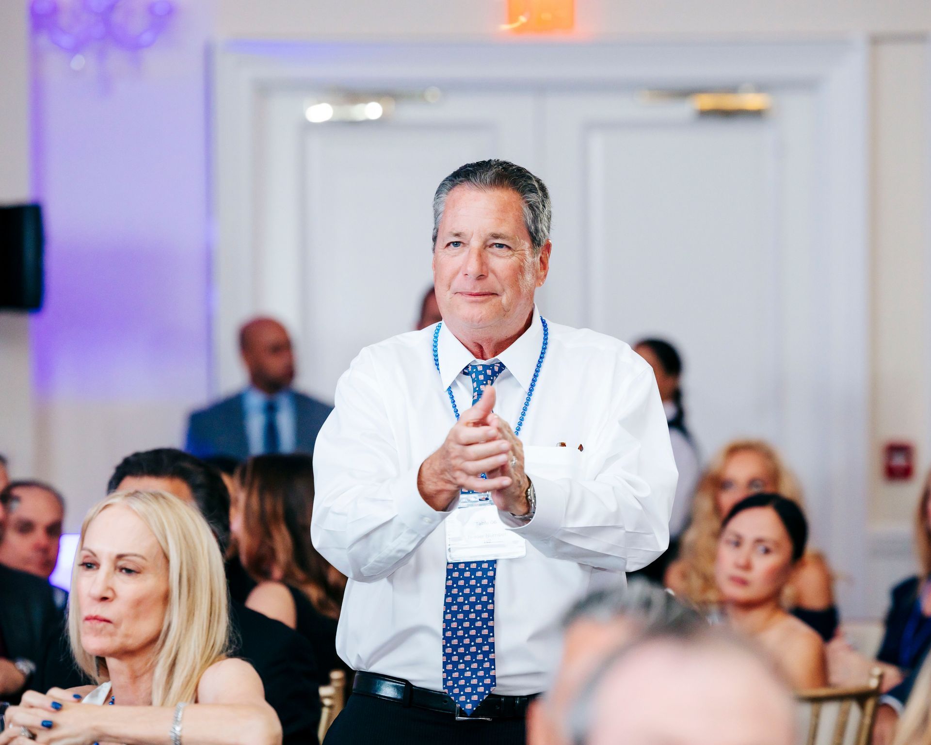 Man in white shirt applauding at event, people in background.