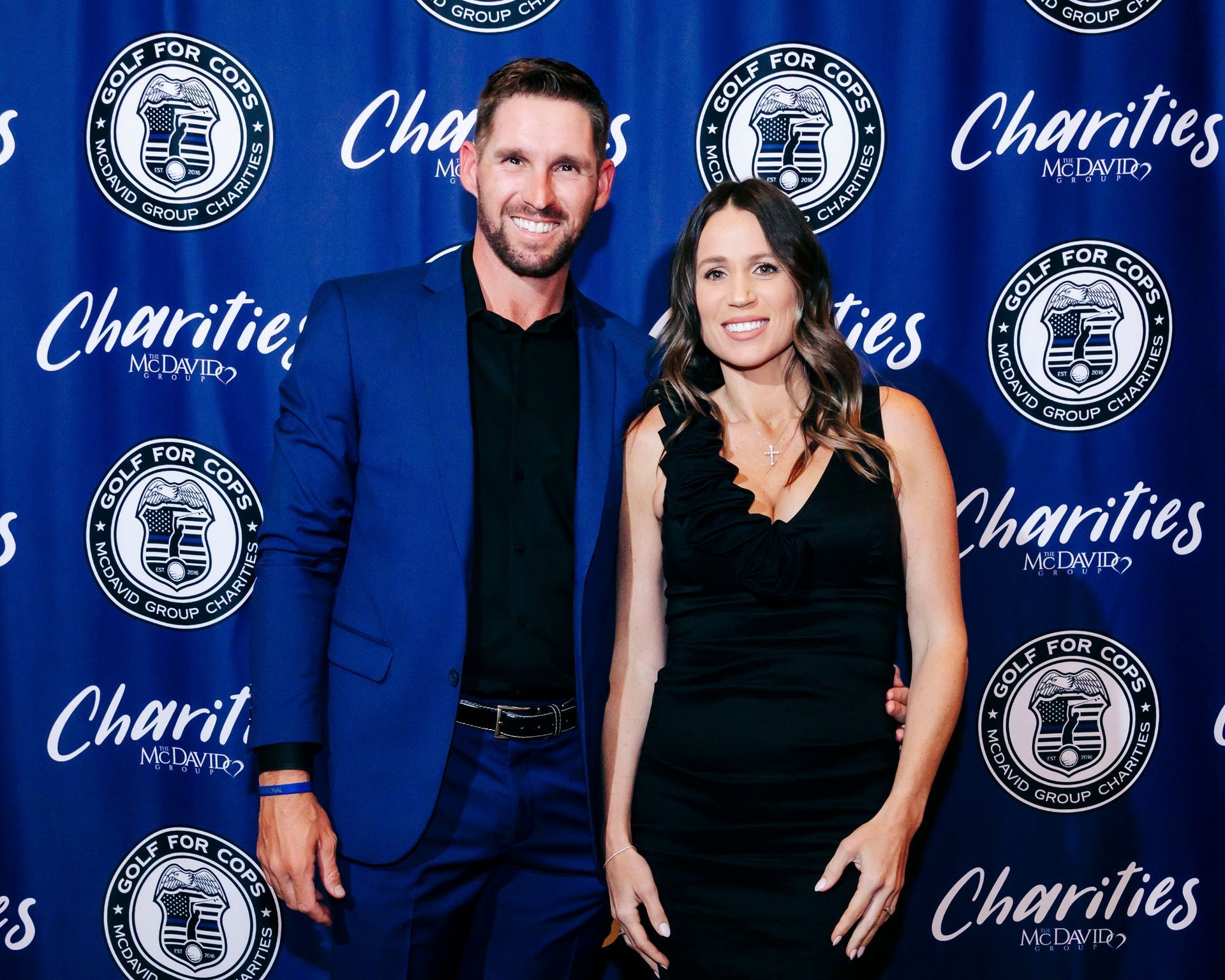 Man and woman posing in front of a blue backdrop with logos. Man in blue suit, woman in black dress.