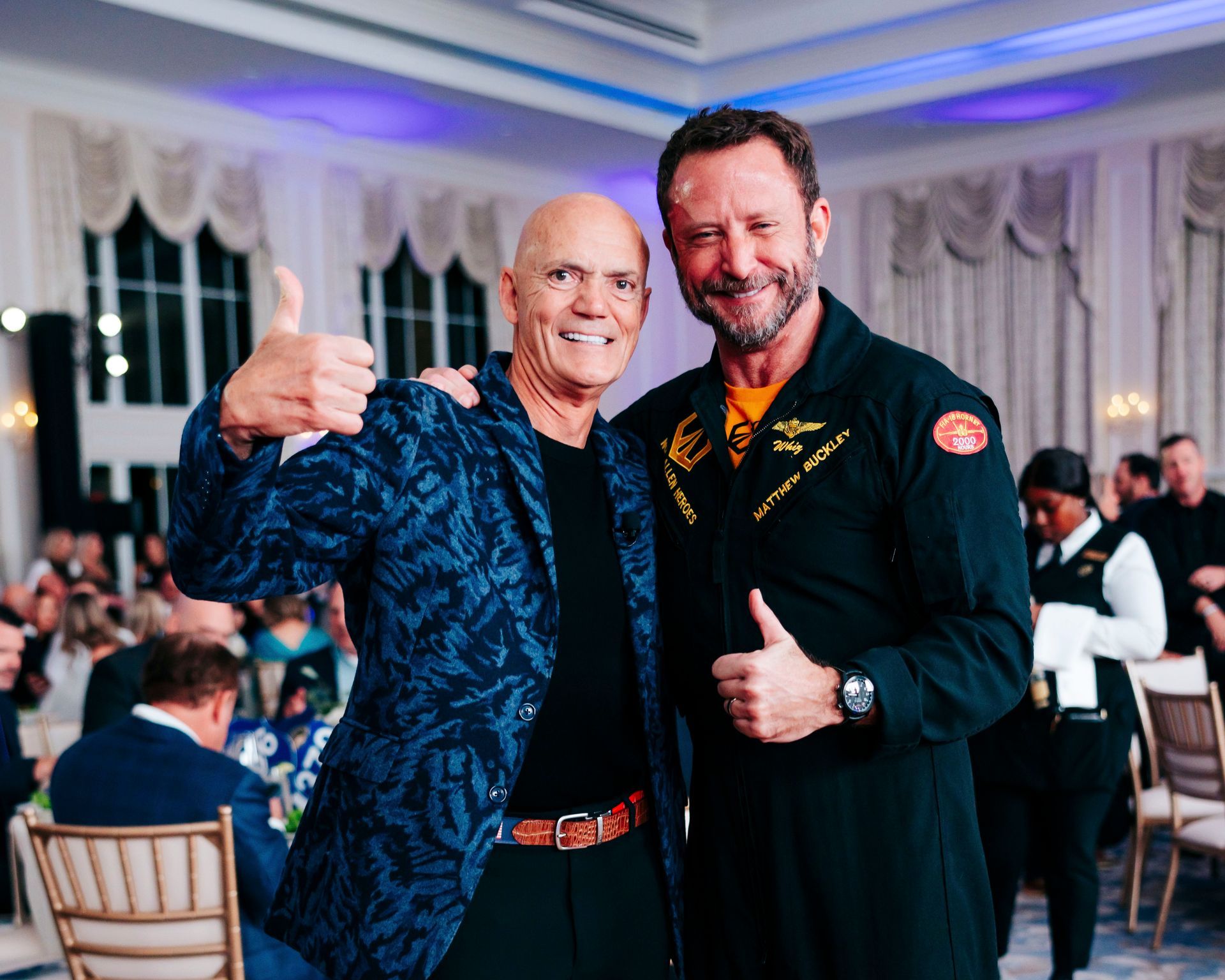Two men smiling and giving thumbs up at an event, one in a patterned jacket and one in a flight suit.