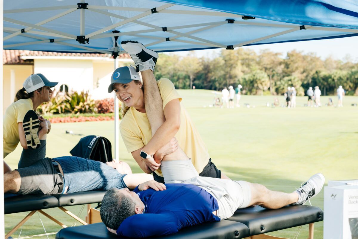 Two people stretch legs on massage tables under a canopy, one assisting the other. Green grass outdoors.