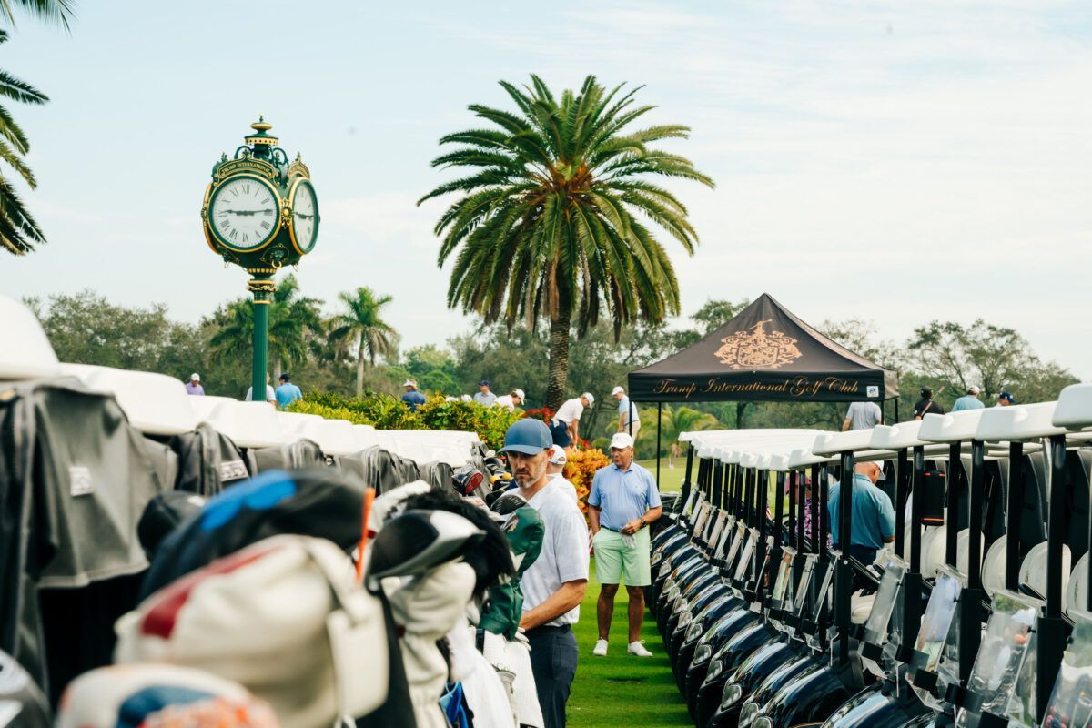 Golfers near carts and golf bags in a sunny, outdoor setting, with a clock tower and palm trees.
