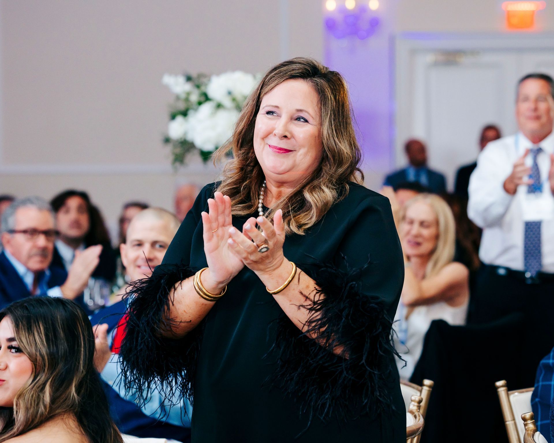 Woman in black dress applauds at event. Attendees in background.
