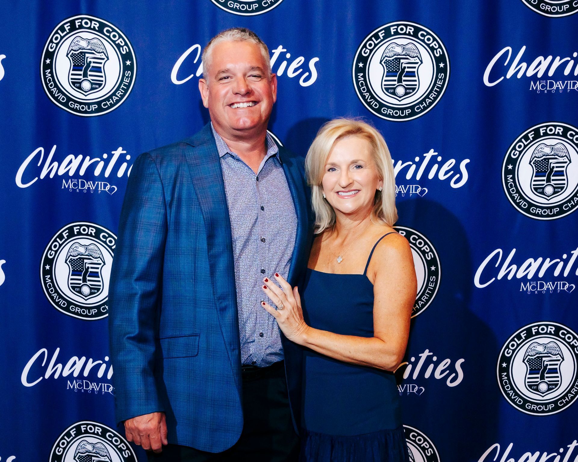 A couple poses in front of a blue backdrop with a logo. Man wears a blue blazer. Woman in a navy dress.