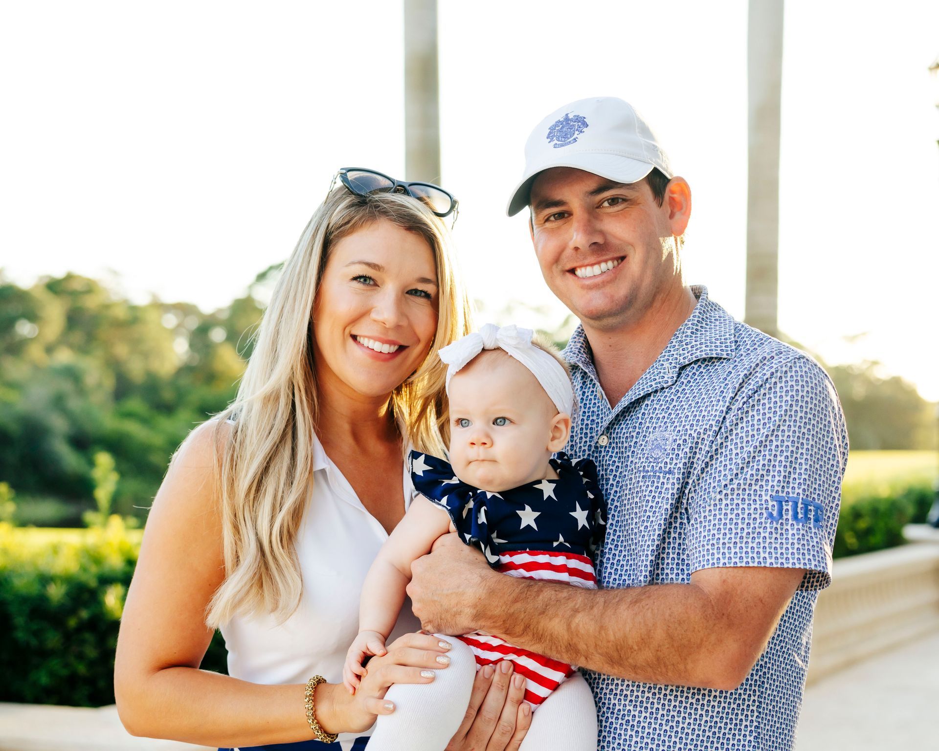 Family of three smiling outdoors: woman with sunglasses, man in cap, and baby in arms.
