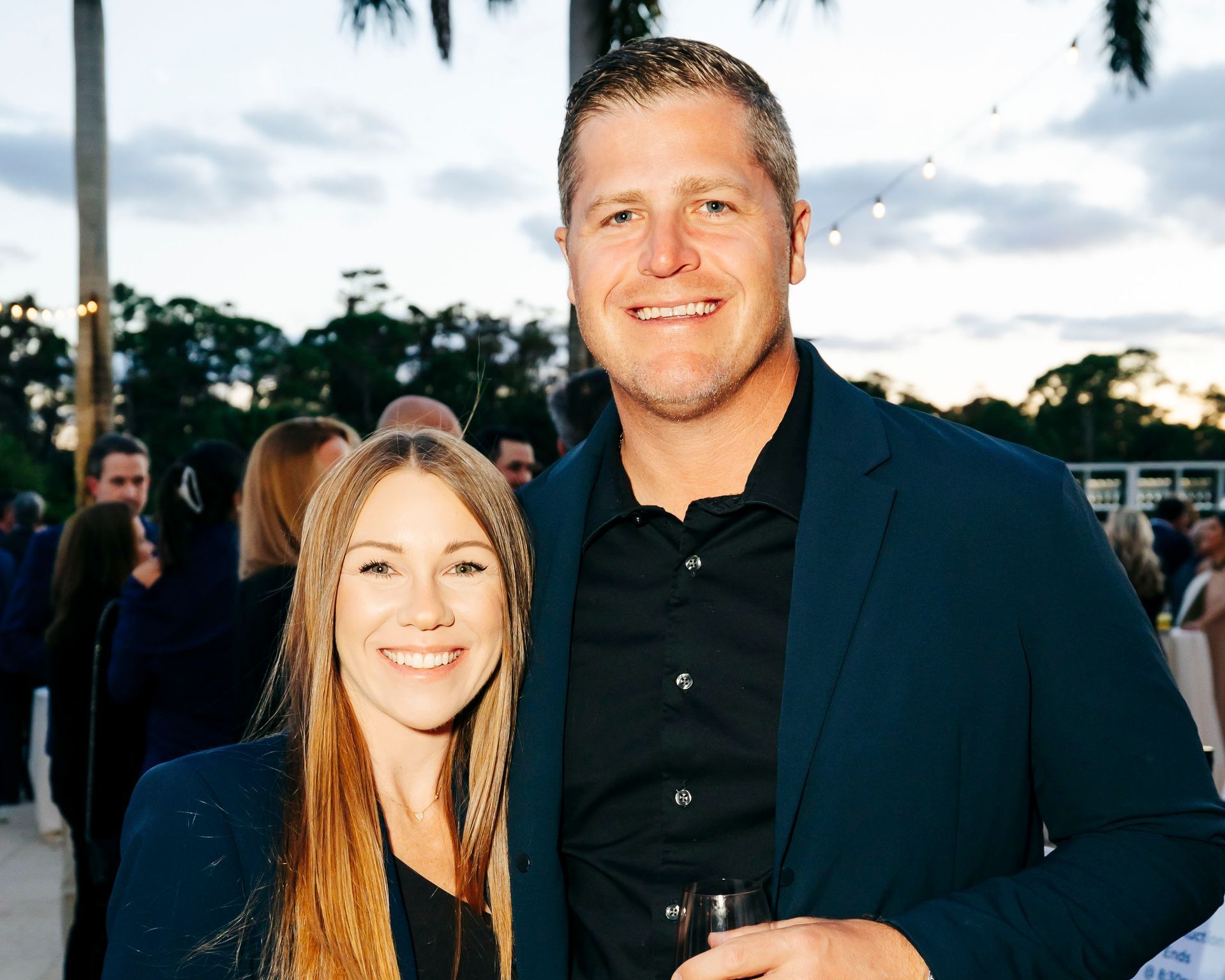 A smiling couple poses outdoors at an event. Woman has long blonde hair; man wears a blue blazer and holds a glass.