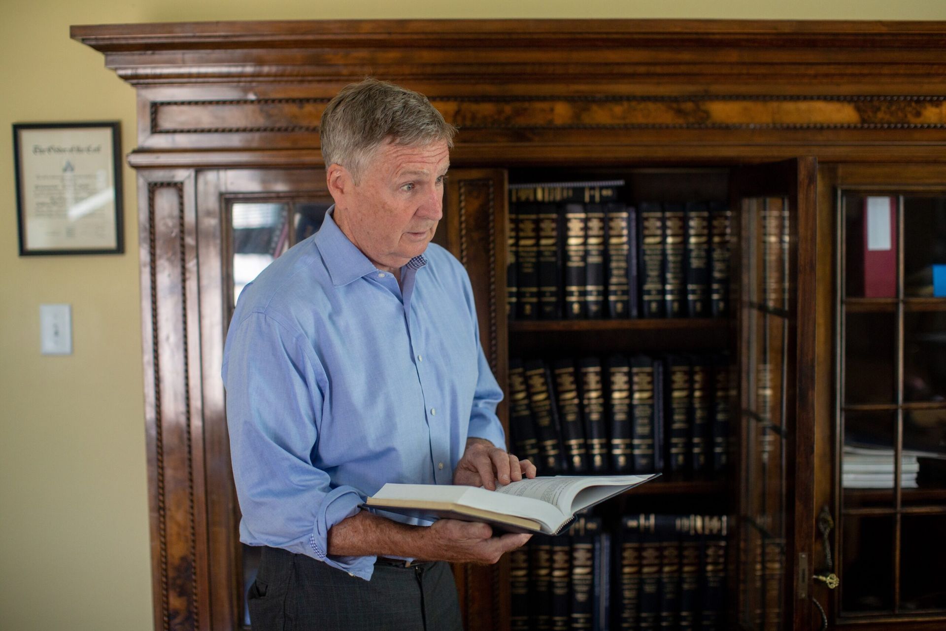 A person wearing a blue shirt reads from an open book while standing in front of a tall wooden bookcase filled with books.