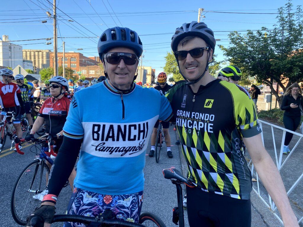 Two cyclists wearing helmets and sunglasses pose together outdoors at a starting line, with other riders in the background.