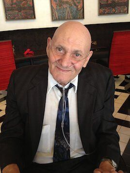 A smiling person with a nasal cannula wearing a suit and tie, sitting in a room with framed artwork and red chairs.