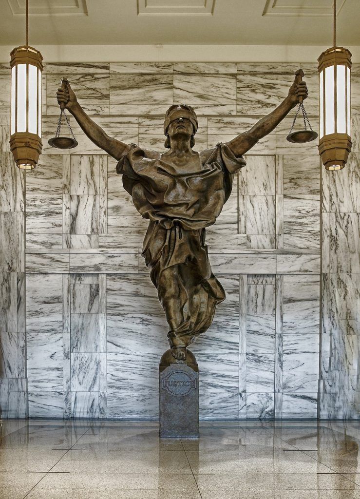 A bronze statue of a figure with outstretched arms holding scales, standing against a patterned white marble wall.