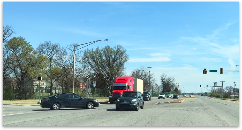 A red semi-truck and several cars driving through a suburban intersection on a sunny day under a clear blue sky.