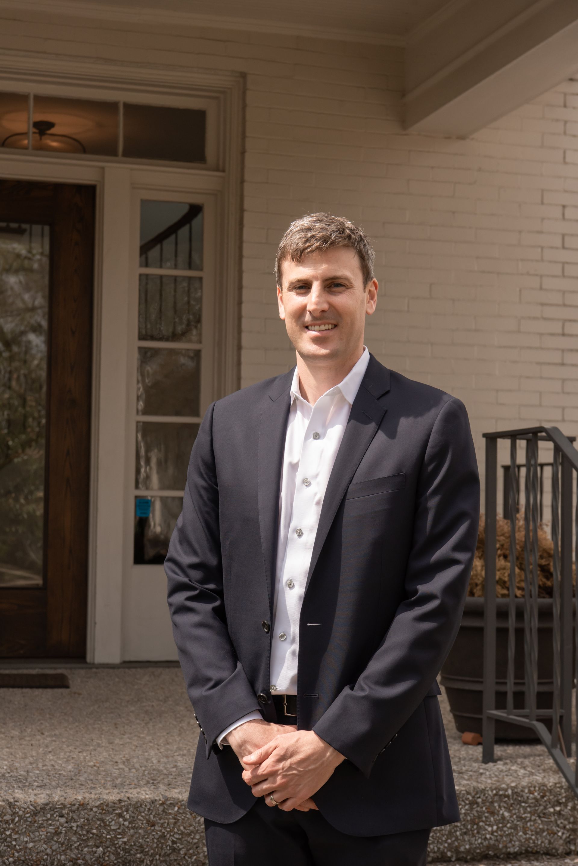 A person in a dark blazer and white button-down shirt stands smiling in front of a house entrance.