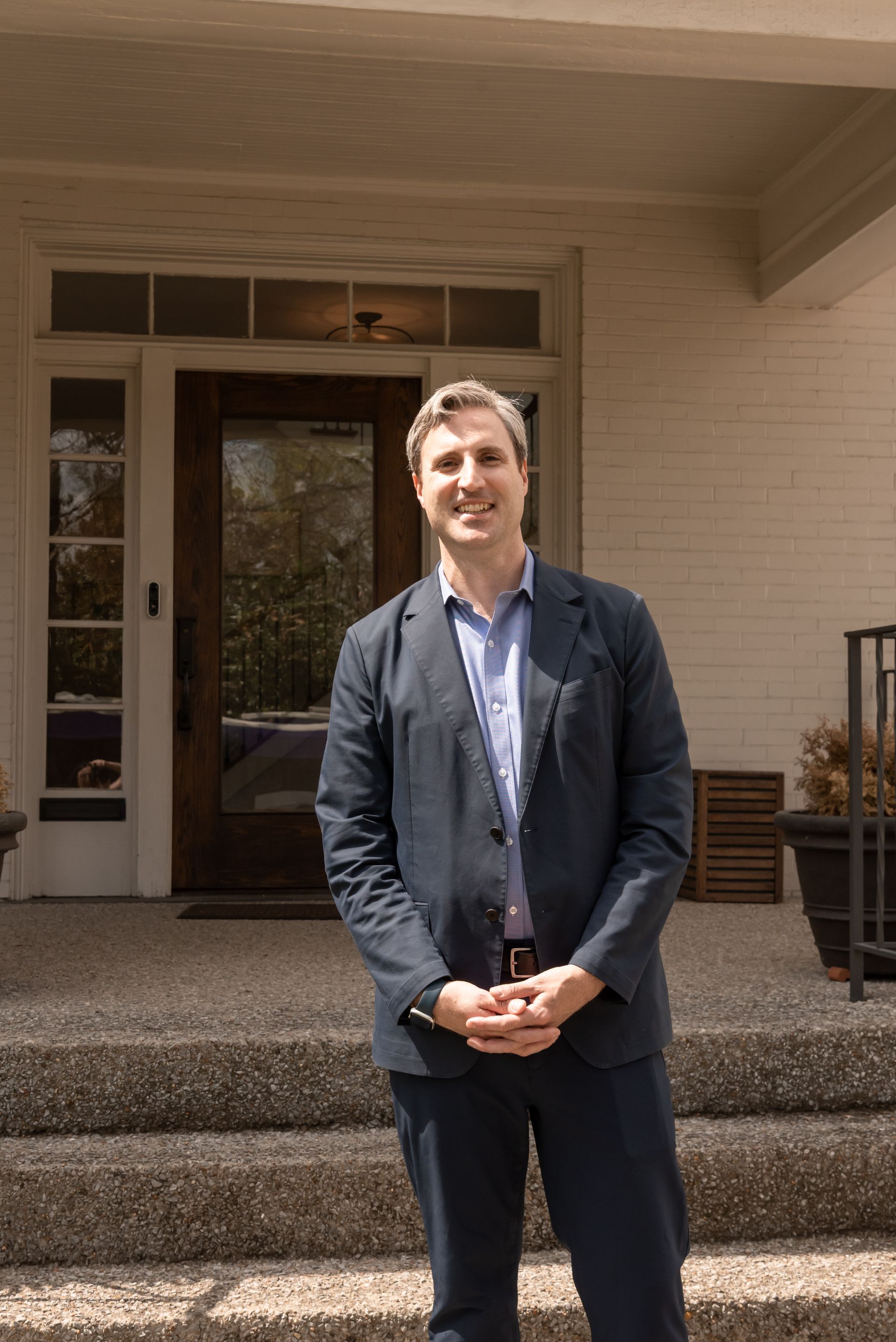A smiling person in a blue blazer and light shirt stands with hands clasped on the steps in front of a house entrance.