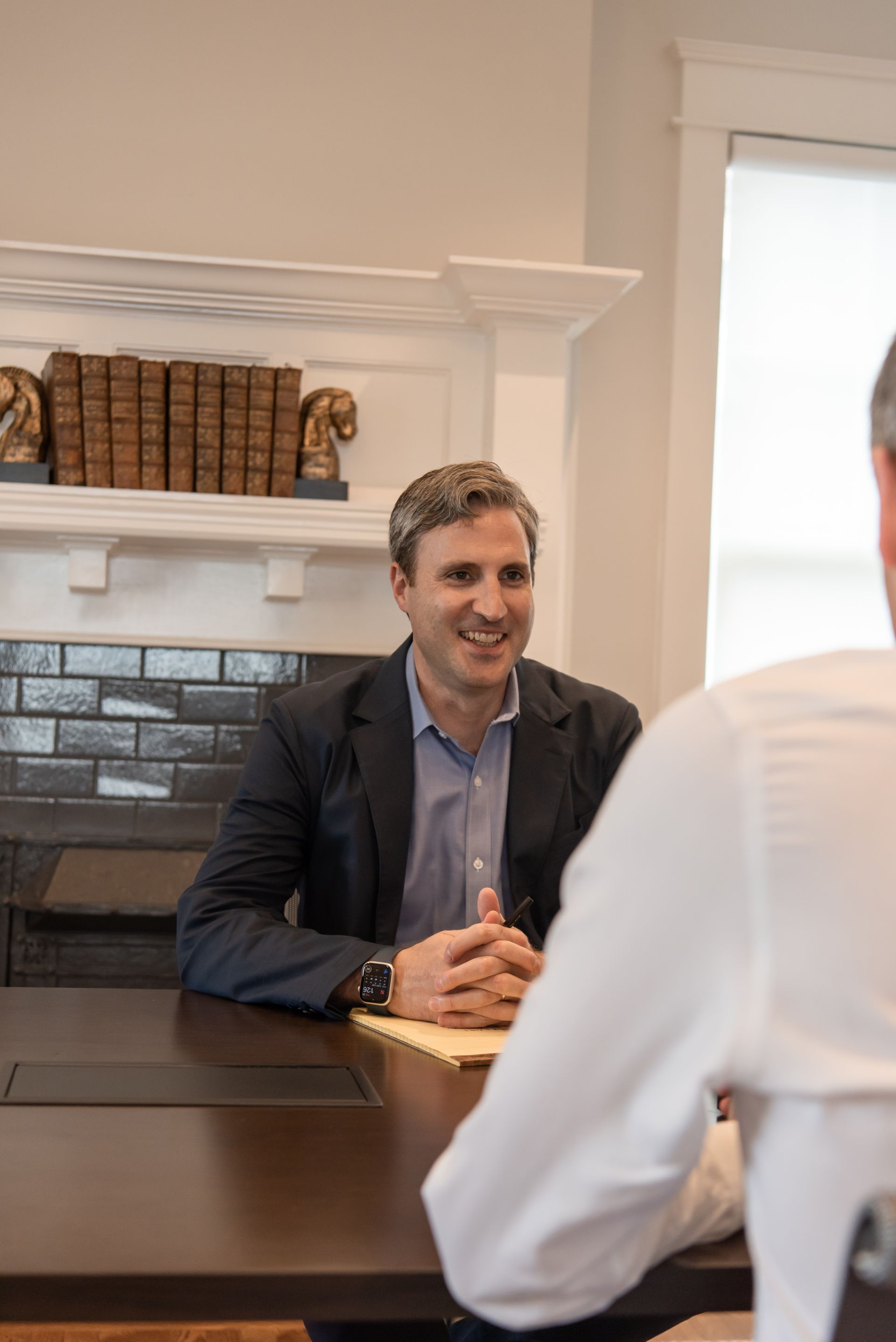 A smiling person in a suit sits at a table across from an individual in a white shirt, inside a bright office.