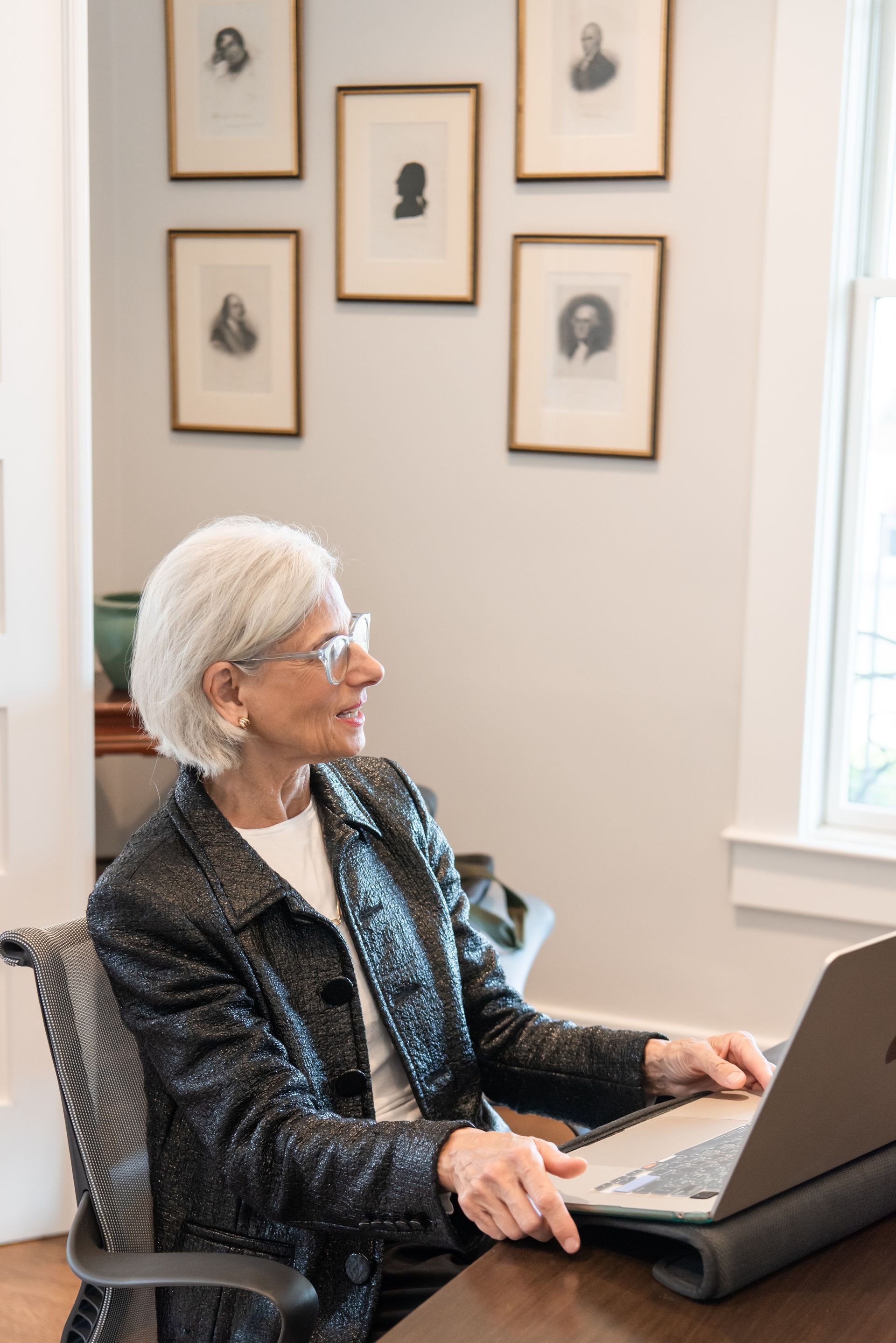 A person with short white hair and glasses wearing a dark jacket uses a laptop while sitting at a desk with framed art.